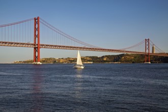 A sailboat passes under a large orange suspension bridge on calm blue waters during evening light,