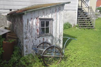 An old, weathered wooden shed with a bicycle trailer in front of it, surrounded by grass and a