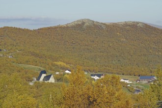 A village with several houses on a hilly terrain, surrounded by autumnal trees and mountains in the