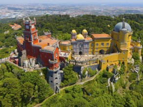 Colourful palace with yellow and red buildings, dome and towers, surrounded by wooded hills, aerial