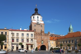 Historic city gate with brick tower and turrets in front of a lively square with passers-by, Krakow