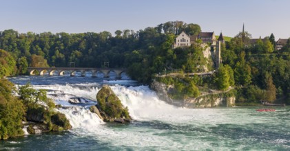 Waterfall flowing next to a historic bridge with lush vegetation and buildings in the background,