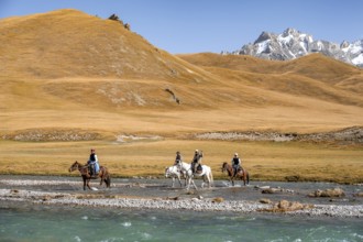 Riders riding through river in front of mountain landscape with yellow meadows, Kol Suu River and