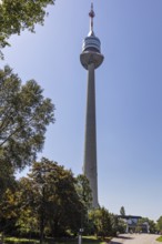 A tall television tower rises into the blue sky above green trees, Vienna