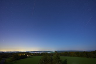 Night scene with starry sky with aeroplane tracks over a quiet, green landscape and trees on the