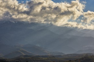 View of hilly landscape with clouds and sun rays shining through the haze, Lefka Ori, White