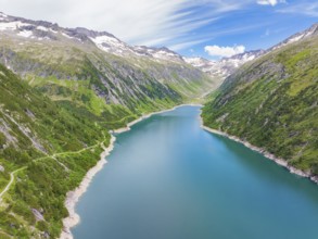 A blue lake meanders through a mountain landscape lined with green hills and snow-capped mountains,