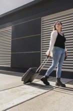 Young business woman pulling rolling suitcase while walking outside airport terminal, preparing to