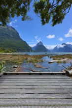 Wooden walkway with a view of mountains and water, framed by lush vegetation under a blue sky,