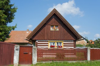 Brown wooden house with a gable and red shutters, surrounded by a wooden fence and flowers, Old