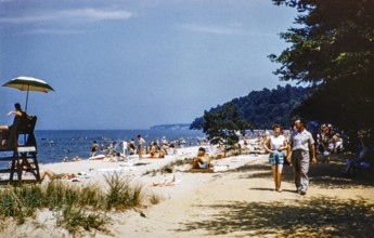 People enjoying the summer sun on a crowded beach with a lifeguard standing guard, USA around 1953