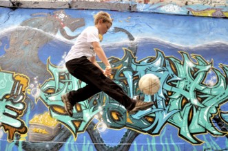 A ten-year-old boy plays with his football in front of a graffiti wall, football pitch in Germany