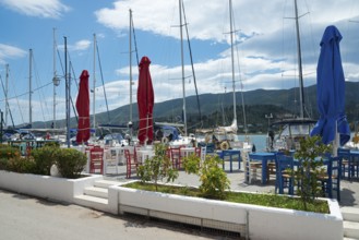 A harbour with moored boats and empty outdoor tables in front of mountains and a cloudy sky, Poros,