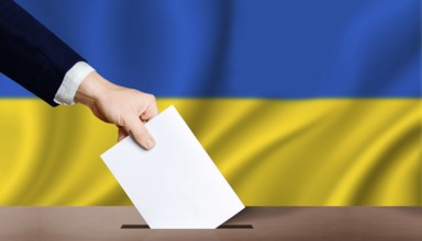 Hand holding ballot in voting ballot box with ukraine flag in background. People puts ballot paper