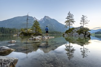 Young man standing on stones in the lake, Hochkalter reflected in Hintersee, at sunset,