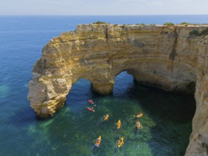 A rocky cliff juts out into the turquoise sea, where several colourful kayaks paddle on the water,