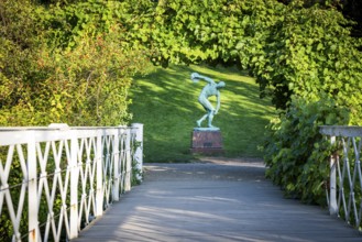 Wooden bridge leading to the sculpture of a discus launcher in a green, sunny park area, Botanical