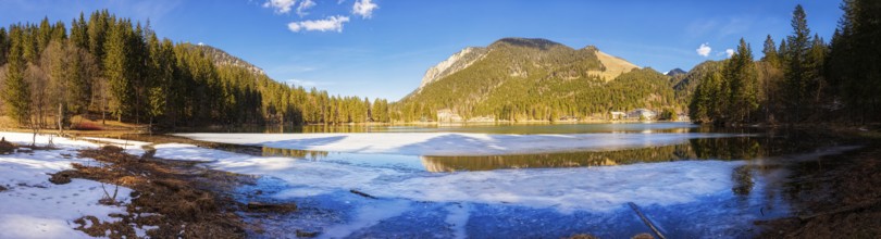 Mountain lake partly covered with snow, surrounded by forests and mountains under a blue sky,