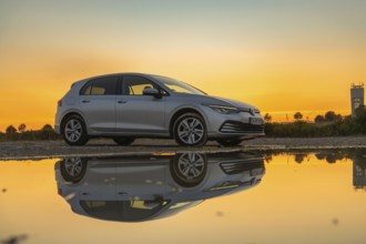 Silver car in setting sun, perfectly reflected in puddle, strong colours in the sky, VW Golff