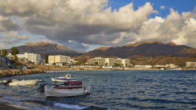 Coastal landscape with calm water, docked boats and mountains in the background, under a cloudy