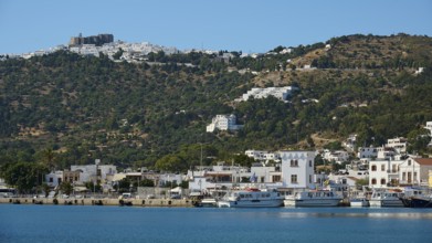 Coastal town with harbour, surrounded by white buildings, at the base of green hills and blue sky,