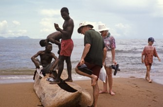 Tourists with local young men looking at a dugout tree on the beach of Lake Malawi, Malawi,