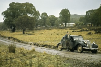 Young man Standing by Citrön Traction Avant car on unmade rural road, Victoria, Australia 1956