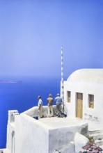 Three male men tourists look out to sea from terrace of whitewashed building at Oia, Santorini,