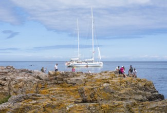 People on rocks by the sea with sailboat in the background in Gudhjem, Bornholm, Baltic Sea,