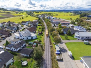 Aerial view of a train on the track through a green residential area with a view of fields and