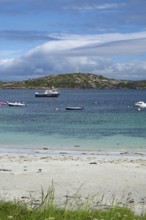 Boats in blue water in front of an island, with white clouds and a beach in the foreground, Holy