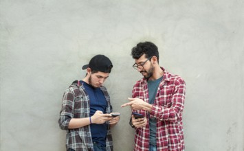 Two young friends leaning against a wall checking their cell phones, Two friends leaning against a