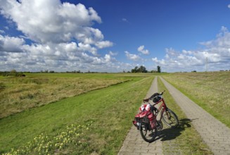 A bicycle stands on a paved path leading through a vast meadow landscape under a cloudy sky, dyke,