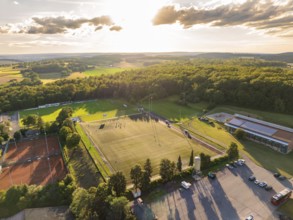 Aerial view of a sports field with football pitch and tennis courts, surrounded by forest and