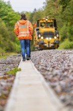 A man in an orange jacket walks along a railway line, a yellow tractor follows, track construction