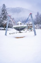 A snow-covered playground equipment on a snowy square, surrounded by fog-covered trees,