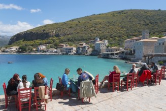 People sitting on red chairs by the sea in front of a picturesque coastal town, Limeni, Areopoli,