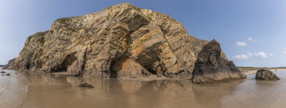 A beach with a rocky cliff in the background. The water is calm and the reflection of the cliff is