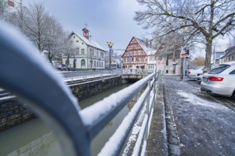 Road with snow cover next to river, view of half-timbered houses and bridge in wintry cityscape,