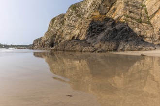 A beach with a rocky cliff in the background. The water is calm and the reflection of the cliff is