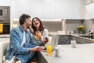 Young couple laughing while using laptop and drinking orange juice in their modern kitchen