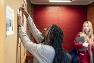 Focused student pinning a paper on a bulletin board in a university hallway, with another student