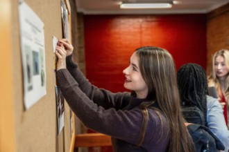 Female college student pinning an important notice on a bulletin board in a busy campus hallway,