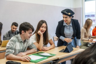 University teacher using tablet explaining lesson to two students sitting at desk in classroom