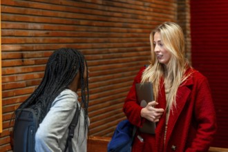 Two female students chatting during break time in university corridor, holding laptop and backpack