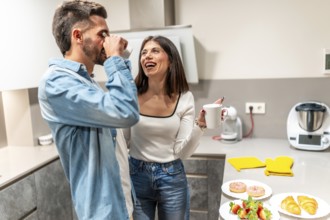 Happy couple drinking coffee and laughing together while having breakfast in their modern kitchen