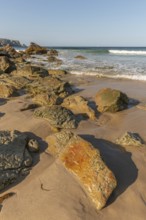 A rocky beach with a large rock in the foreground and a boat in the distance. The beach is quiet