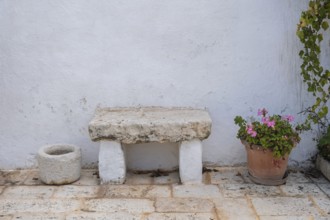 Stone bench and flower pot with geraniums on a rustic stone floor in front of a white wall, Apulia,