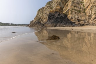 A beach with a rocky cliff in the background. The water is calm and the reflection of the cliff is