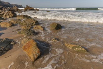 A rocky beach with a large rock in the foreground and a boat in the distance. The beach is quiet